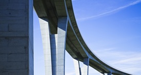 Ein modernes Brückenbauwerk mit hohen, schlanken Betonpfeilern ragt vor blauem Himmel empor. Die Brücke schwingt elegant in einer Kurve und vermittelt Stabilität sowie technische Präzision.

                                    